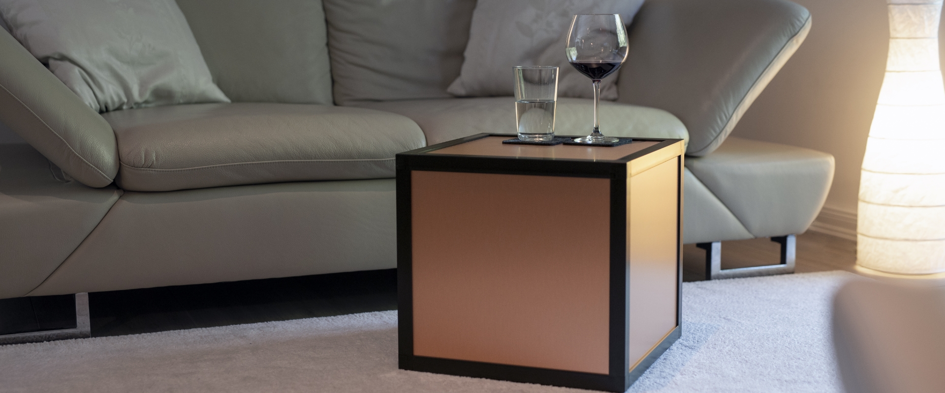 Eine würfelförmige boxula mit Rahmenfarbe Rich Black und Seitenwänden in Copper brushed steht auf einem Teppich in einem Wohnzimmer, nebenan befindet sich ein Sofa. Auf der boxula stehen ein Glas mit Wasser und ein befülltes Weinglas.